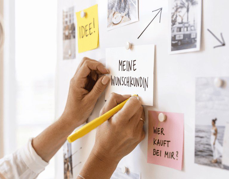 Hände ordnen Notizzettel und Fotos zur Wunschkundin auf einer hellen Pinnwand mit gelbem Marker